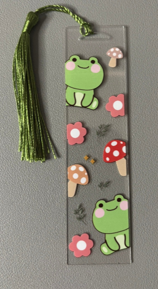 Bookmark with green frog, pink flowers, and red-spotted mushrooms on a gray background