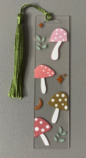 Decorative bookmark with colorful mushroom designs and a green tassel on a gray background